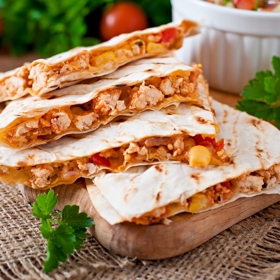 Stacked chicken and cheese quesadillas with corn and peppers, garnished with parsley on a wooden board.