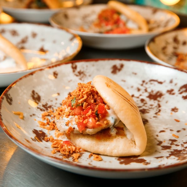 A Mexican bao bun with crispy onions, diced tomatoes, and white sauce on a decorative plate.